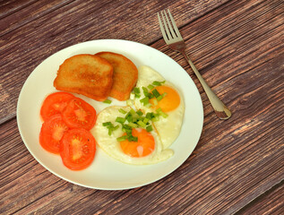 Delicious breakfast, plate with two fried eggs, toasts of wheat bread and pieces of ripe tomato on wooden table.