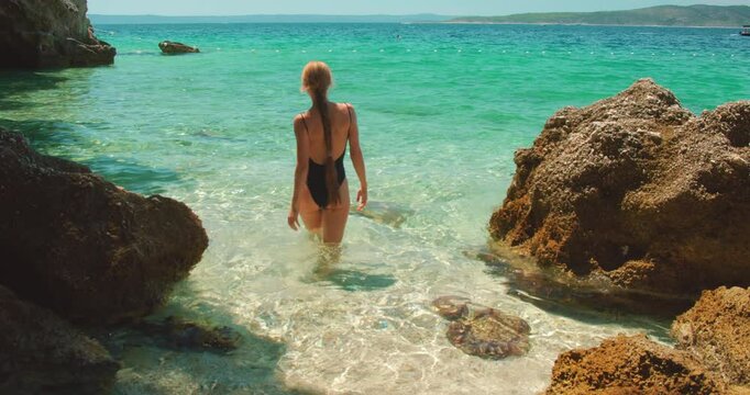 Woman walks into shallow emerald sea water between rocks on sunny day in Croatia