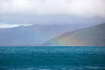 A faint rainbow arches from a cloudy sky onto a blue wind swept ocean adjacent a tropical coastal...