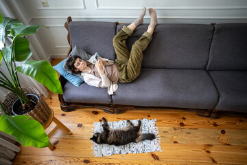 Indifferent woman lounging in lazy pose scrolling through smartphone social media, cat rests close...