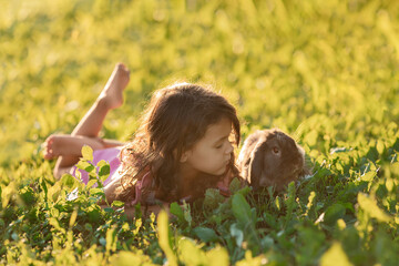 Toddler girl lying on green grass and playing with brown rabbit at summer days