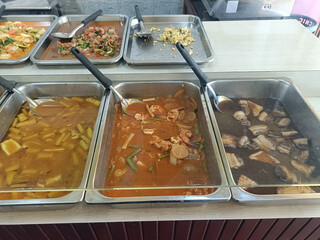 Variety of hot dishes in stainless steel trays with serving spoons, including curry, stew, and...