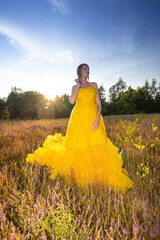 A beautiful woman in a yellow dress stands gracefully in a sunlit field during the enchanting golden hour