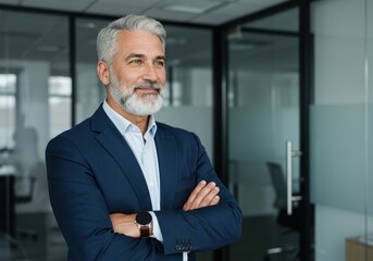Confident senior businessman with gray hair and beard standing with arms crossed in modern office