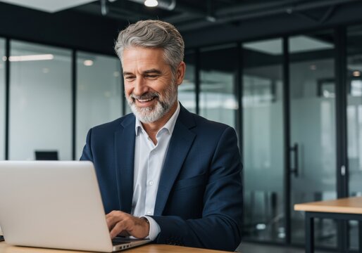 Smiling senior businessman in a navy suit actively typing on a laptop in a modern office environment - Powered by Adobe