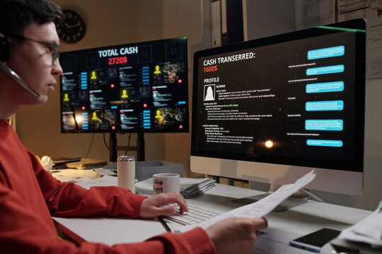 Young man wearing headset operating computer in scam call center, analyzing victim profiles and transaction data on multiple screens
