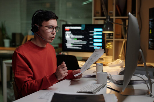 Young man wearing headset sitting at desk holding documents and talking while working at computer with multiple monitors in scam call center - Powered by Adobe