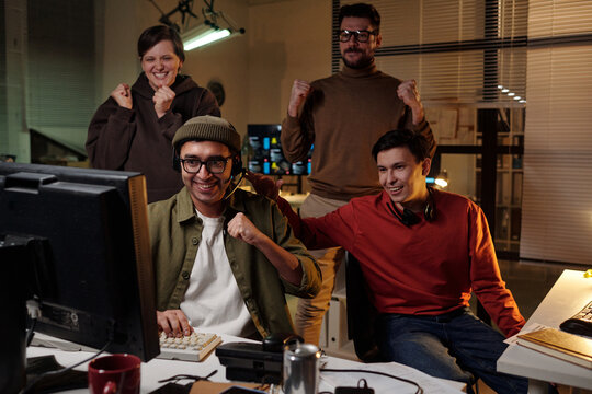Group of young adult multiethnic men and woman celebrating while sitting and standing around computer monitors in scam call center - Powered by Adobe