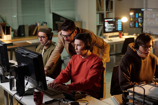 Group of people working at computers in call center, wearing headsets, concentrating on screens, supervisor standing behind, scam call center context