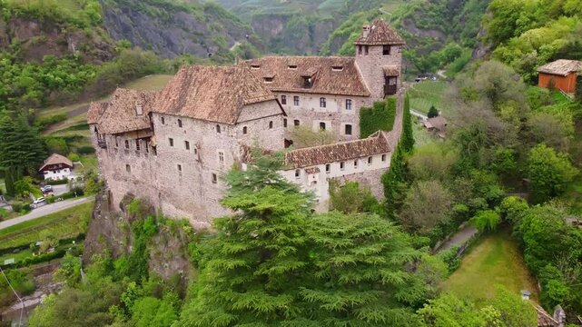 Drone flying toward Castel Roncolo (Runkelstein Castle) in Bolzano, Italy, perched on a rocky spur above a valley, surrounded by trees and alpine hills during spring, capturing medieval architecture
