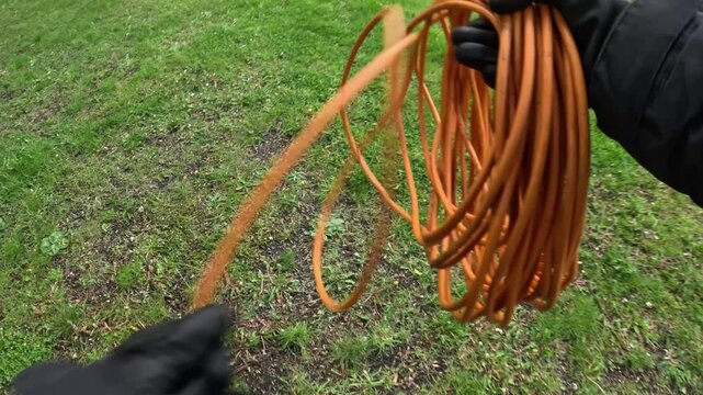 Technician unrolls bright orange power extension cable over grassy outdoor area, captured from a first-person perspective with gloved hands, real time utility prep for event setup, extreme wide angle