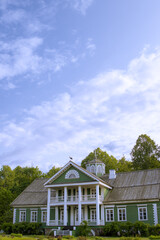 Elegant vintage estate with traditional architecture, white porch pillars, and green painted walls. Nestled in nature and backed by forest trees under a serene, partly cloudy sky. Stork bird on a roof