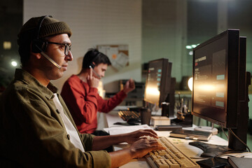 Man wearing headset operating computer in foreground, another man using phone and typing at desk in...