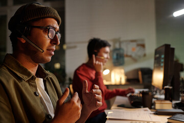 Indian man wearing headset operating computer and gesturing while sitting beside young adult man...