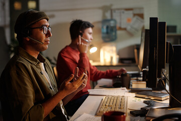 Indian man wearing headset gesturing while speaking at computer, another man in background talking on phone, both working in scam call center at night