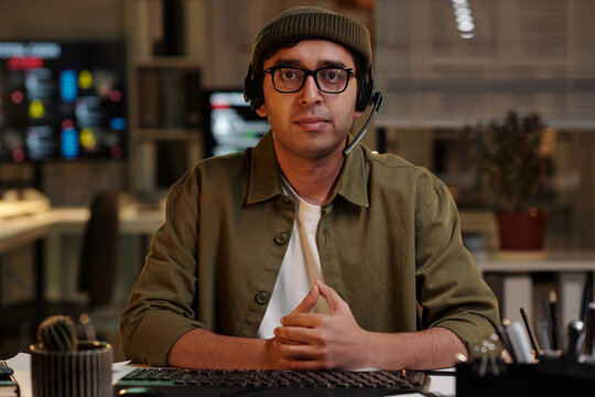 Portrait of Indian man wearing headset sitting at desk in modern office setting, looking directly at camera, hands clasped