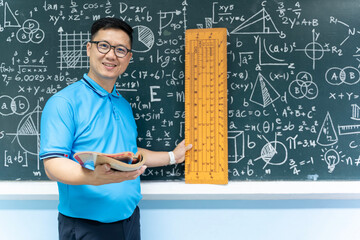 Asian male math teacher holding book and large ruler standing in front of chalkboard with formulas...