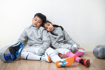 Fencer boy and girl sit on the floor with their backs against a wall, exhausted after a practice or competition.