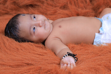 Cute Indian baby lying on a soft textured brown rug, wearing a diaper and traditional black thread...