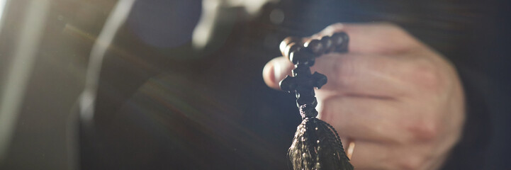 Header of hands of man holding rosary beads inside church, fingers gently gripping wooden cross,...