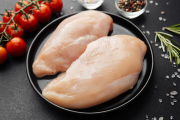 Raw chicken breasts on black plate with tomatoes and spices on dark background