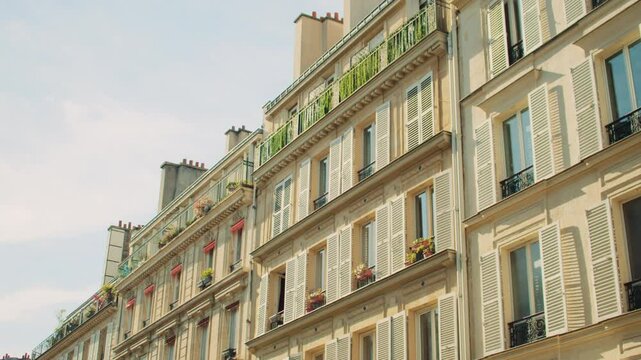Charming Haussmann-style residential buildings in Paris with white shutters, wrought iron balconies, and blooming window plants under bright summer sky. Overview of classic Parisian apartment facades