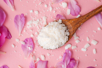 Soy wax flakes on wooden spoon with pink petals on pastel background