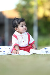 Adorable baby boy sitting on a grassy field with soft printed cushions, wearing a white and red...