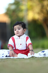 Adorable baby boy sitting on a grassy field with soft printed cushions, wearing a white and red...