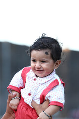 Adorable baby boy sitting on a grassy field with soft printed cushions, wearing a white and red...