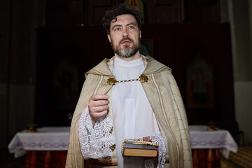 Caucasian middle aged man wearing priest vestments standing in church holding rosary and book,...