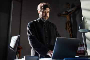 Middle aged Caucasian man with beard wearing clerical robe standing in church using laptop, focused...