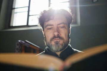 Portrait of middle aged Caucasian man reading large book in church, sunlight streaming through...
