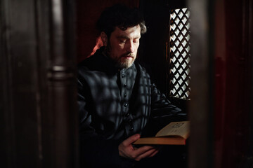 Middle aged man with beard sitting in church confessional reading book, wearing clerical clothing,...
