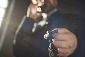 Middle aged man holding rosary beads in hand, standing in church, wearing clerical clothing, face...