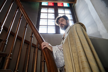 Portrait of middle aged man wearing priest vestments standing on staircase in church, looking into...
