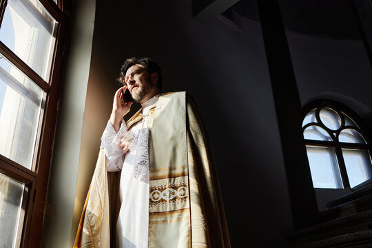 Middle aged Caucasian man wearing priest vestments standing near church window talking on smartphone, sunlight illuminating face, holding hand to chest, looking thoughtful