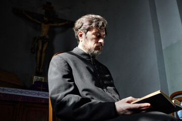 Middle aged Caucasian man sitting in church reading book, wearing clerical robe, head slightly...