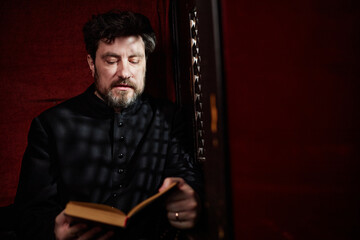 Middle aged Caucasian man wearing clerical clothing sitting in church reading book with serious...