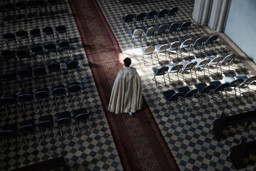 Caucasian middle aged male priest wearing robe walking down church aisle between rows of empty...