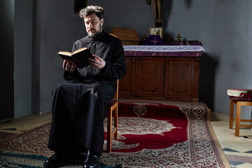 Middle aged Caucasian man wearing clerical robe sitting on chair in church reading book with...