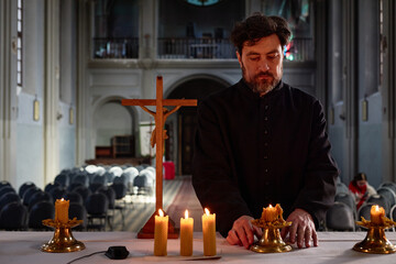 Middle aged man in priest robe arranging candles on altar in church sanctuary, wooden crucifix...