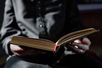 Man sitting in church holding open book with both hands, reading attentively, wearing clerical...