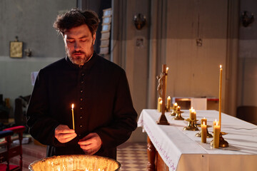 Middle aged Caucasian man wearing clerical robe lighting candle in church, standing near altar with...
