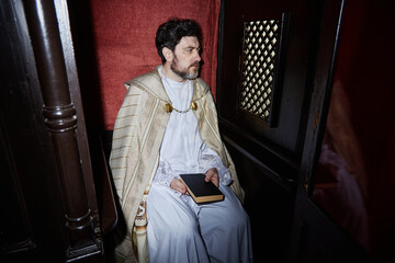 Middle aged man wearing priest robe sitting in church confessional holding book looking to side,...