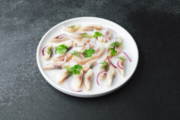 Marinated herring slices with red onion and herbs on white plate over dark background