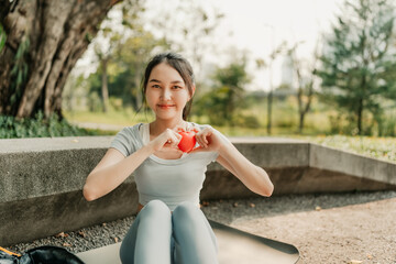 A woman in sportswear sits on a yoga mat in a park, forming a heart shape with her hands and a red...