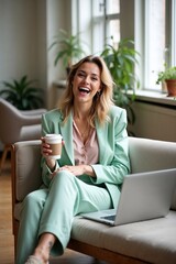 Lachende moderne junge Frau mit To-Go-Kaffeebecher und Laptop auf Sofa in heller Wohnung