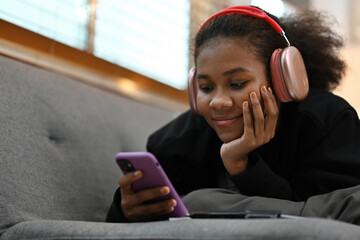 A young African woman in headphones relaxes on the couch, smiling while using her smartphone in a...