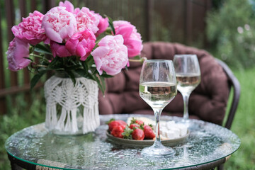 Vase with lush pink peonies, two wine glasses, strawberries and cheese on a glass outdoor table in raindrops. Cozy romantic atmosphere in summer garden